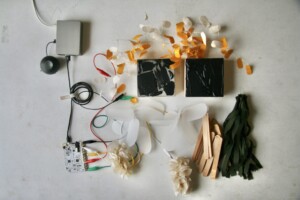 Two small square acrylic paintings on wood are surrounded by miniature mixed media sculptures that are wired to a touch board computer, raspberry pie computer, and a speaker.