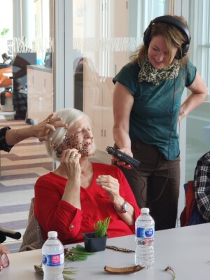 An elderly woman is holding a dried flower to her cheek while Dawn holds a recorder nearby.