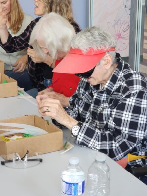 An elderly woman in a red visor and black plaid shirt is tying up collage materials with wire to make a mobile.