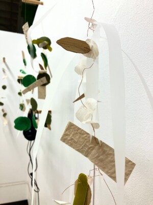 A close up of a mobile sculpture. There are two long white strands resting on top with little pieces of various shapes and beige colours tied down a copper wire.
