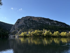 A row of sunlit golden trees along a lake are at the base of a mountain.