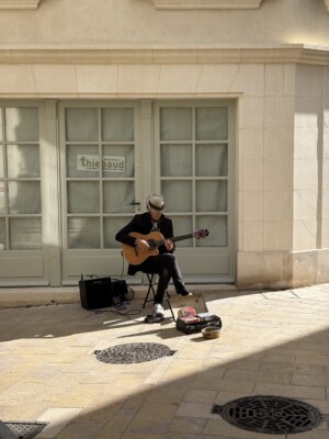A man in black with a tan rimmed hat sits on a chair in a sunlit alley playing a guitar.