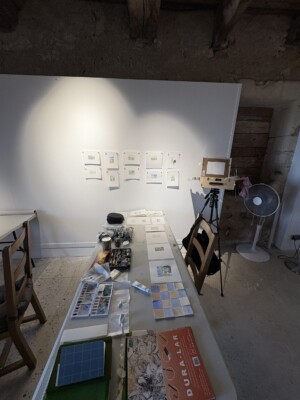 View of my studio desk with supplies and artworks organized on top. Artworks are also shown on the wall above.