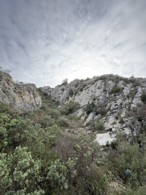 A dramatic gray sky with light shining behind a rocky mountain with shrubs scattered around it.