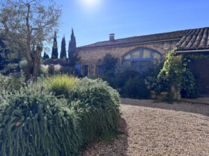 View of the forge building of the moulin among a large mediterranean garden and pebbled driveway in the afternoon light.