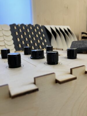 A close up of a DIY synthesizer with a wooden case and seven black knobs. Three small tactile and abstract artworks are displayed behind.