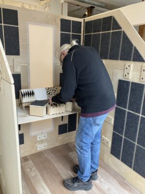 An elder man stands at the table inside the booth playing with the synthesizer knob functions.