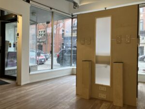 A booth structure with a soft light diffused through a large vertical window and a plinth on either side faces inward inside a gallery with large windows at the back.