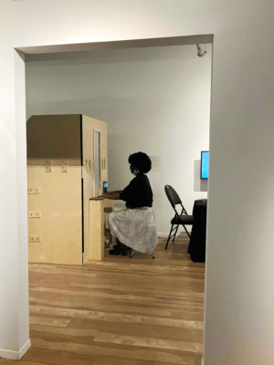 A young black woman is seated in front of the booth with her hands placed on the two plinths in front of her.